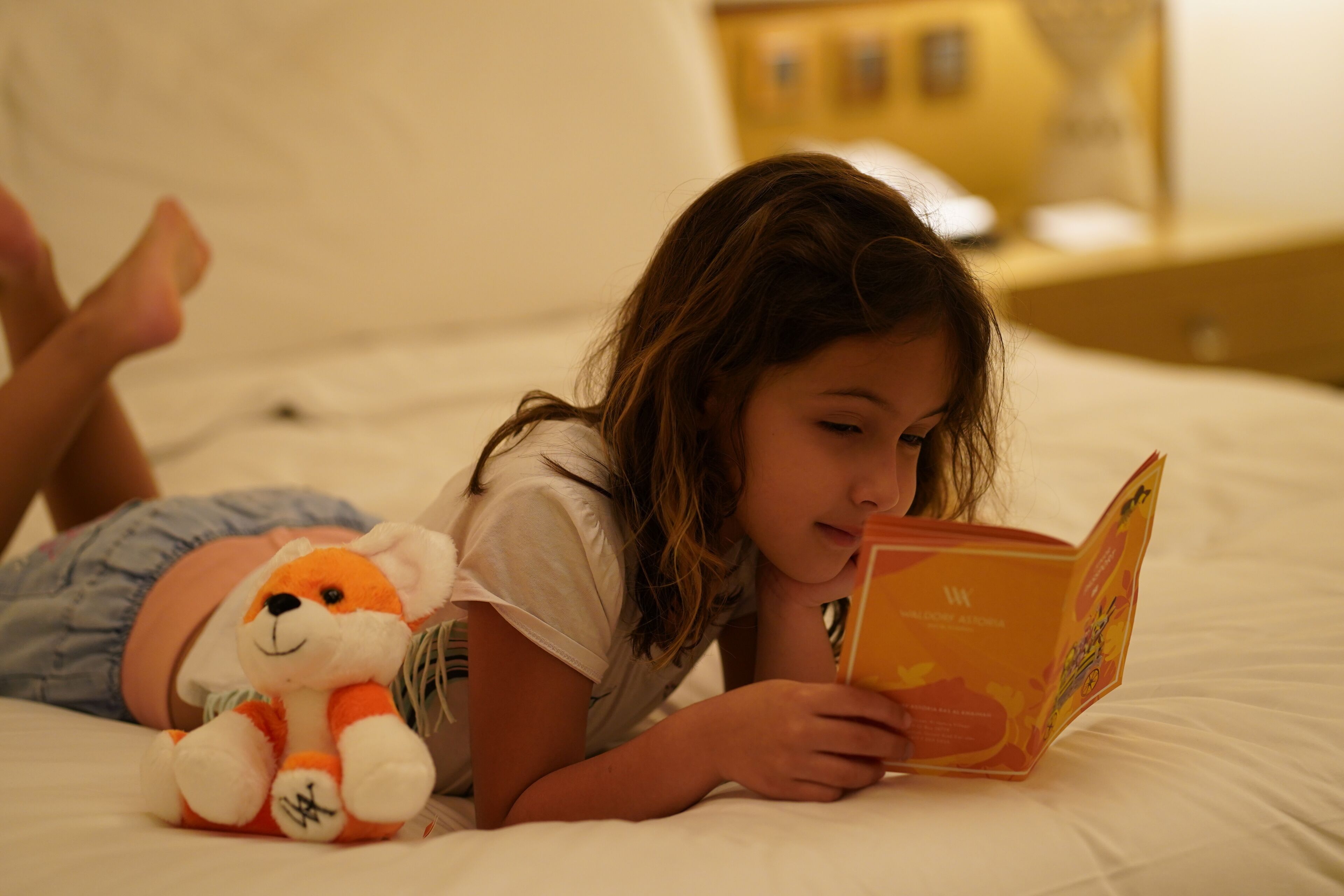 A person lying on a bed reading a book, with a stuffed toy beside them.