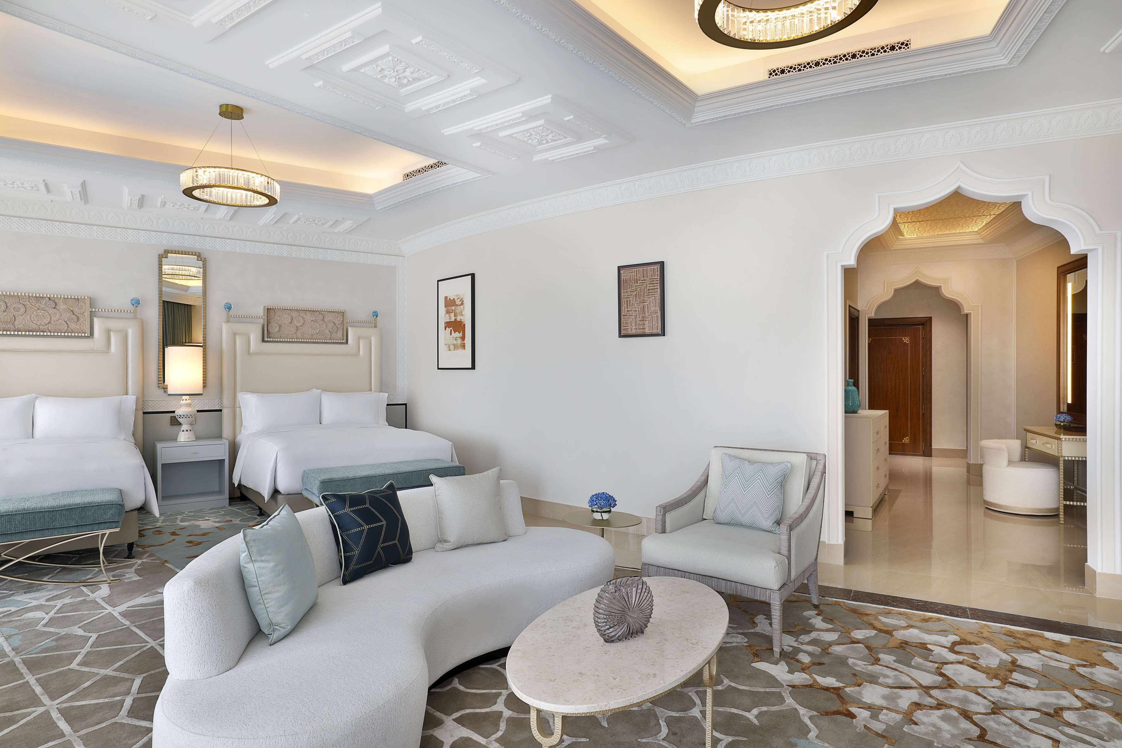 Une chambre d’hôtel avec un canapé incurvé, deux lits simples, un petit fauteuil, une table basse et un lustre.