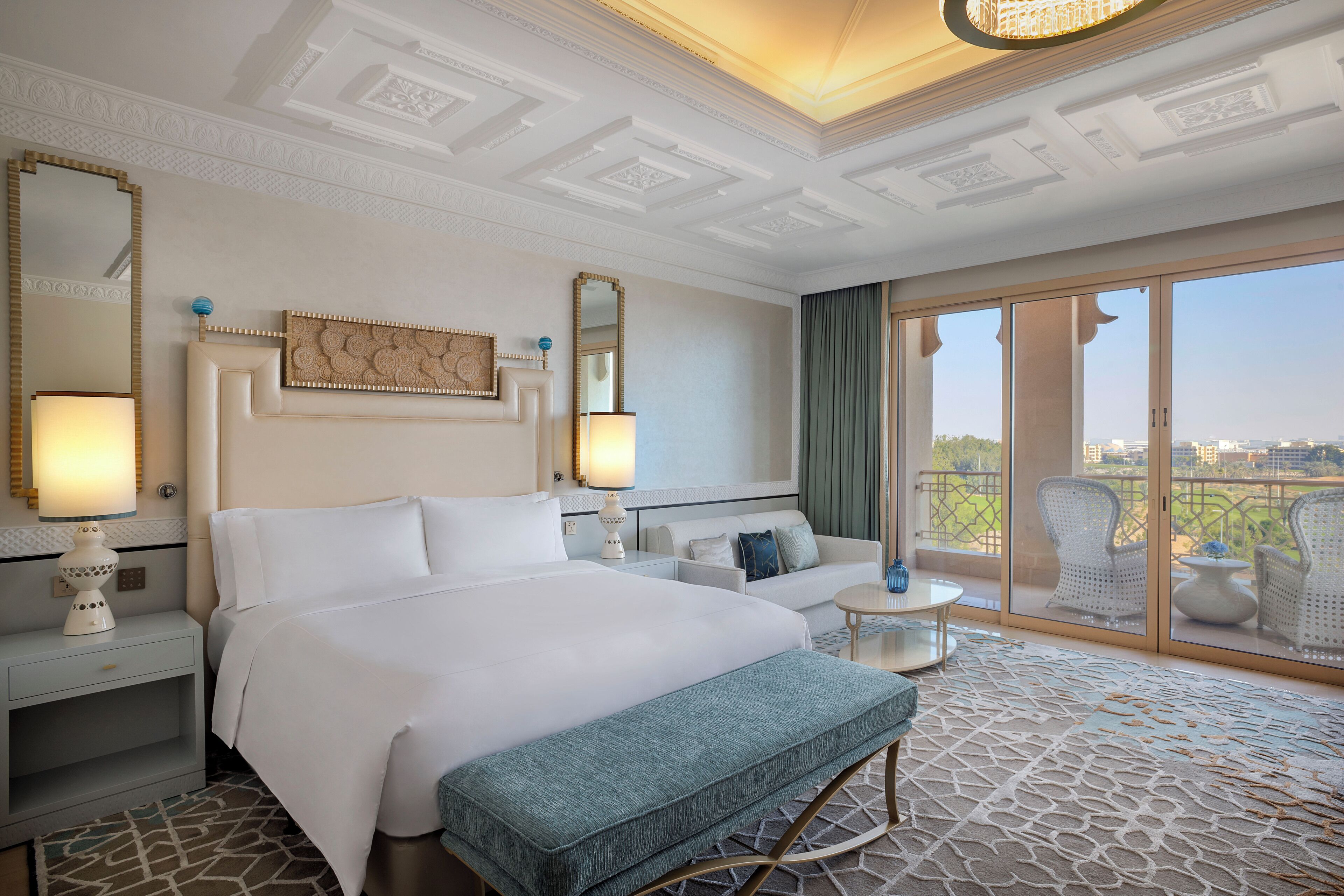 Ein Hotelzimmer mit einem großen Bett, einer Sitzecke mit einer Couch und Sesseln, einem Balkon mit Blick und einer dekorativen Decke.