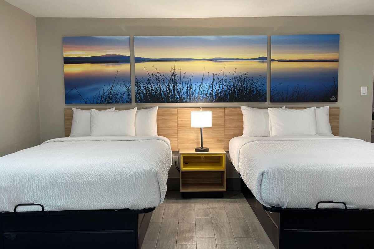 Habitación de hotel con dos camas, una mesita de noche con lámpara y un cuadro grande de paisaje en la pared.