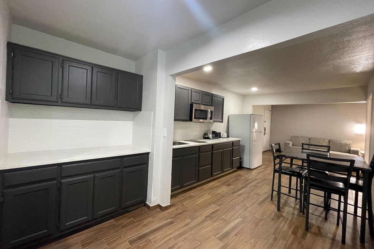 Una cocina moderna con muebles oscuros, un microondas, un refrigerador y una zona de comedor con una mesa y sillas.