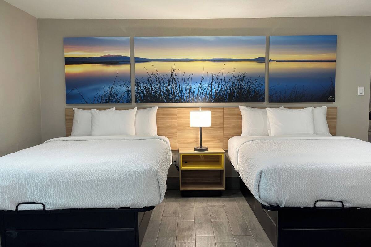 A hotel room with two beds, a nightstand with a lamp, and a large wall painting of a landscape.