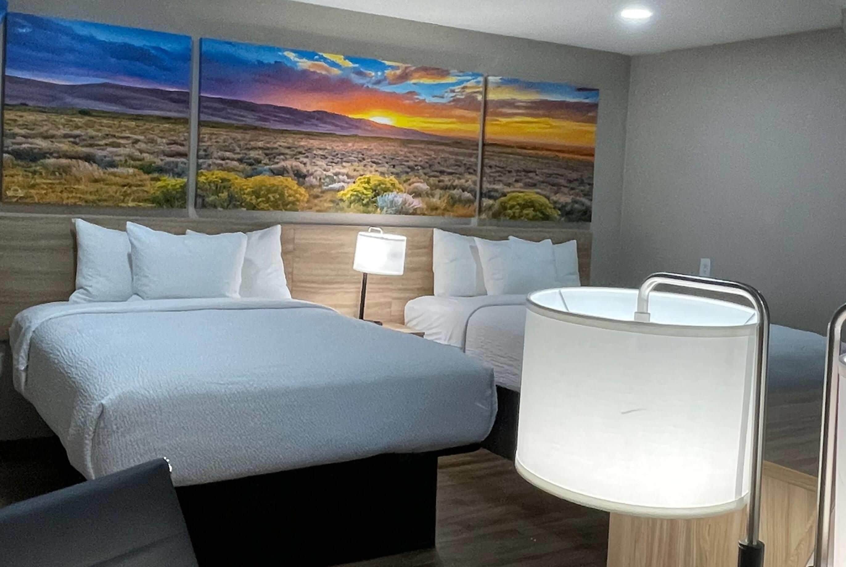 A hotel room with a large bed, a bedside table with a lamp, and a painting of a sunset landscape on the wall.