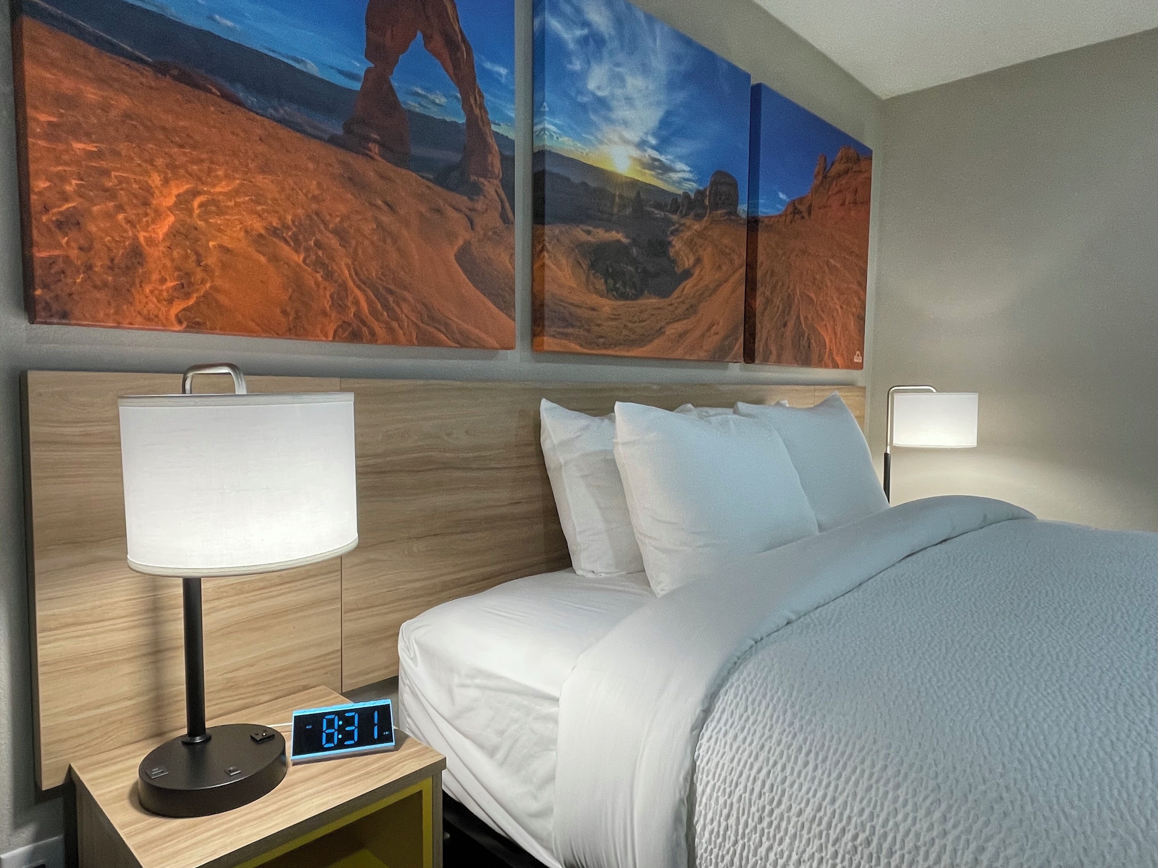 Hotelkamer met een bed, witte beddengoed, nachtkastje met digitale klok, lamp en twee wandlampen.