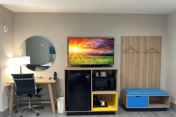 Une chambre d’hôtel compacte comprenant un bureau, un réfrigérateur noir, un four à micro-ondes, un lit bleu, un grand miroir et un téléviseur fixé au mur qui diffuse une scène de coucher de soleil.