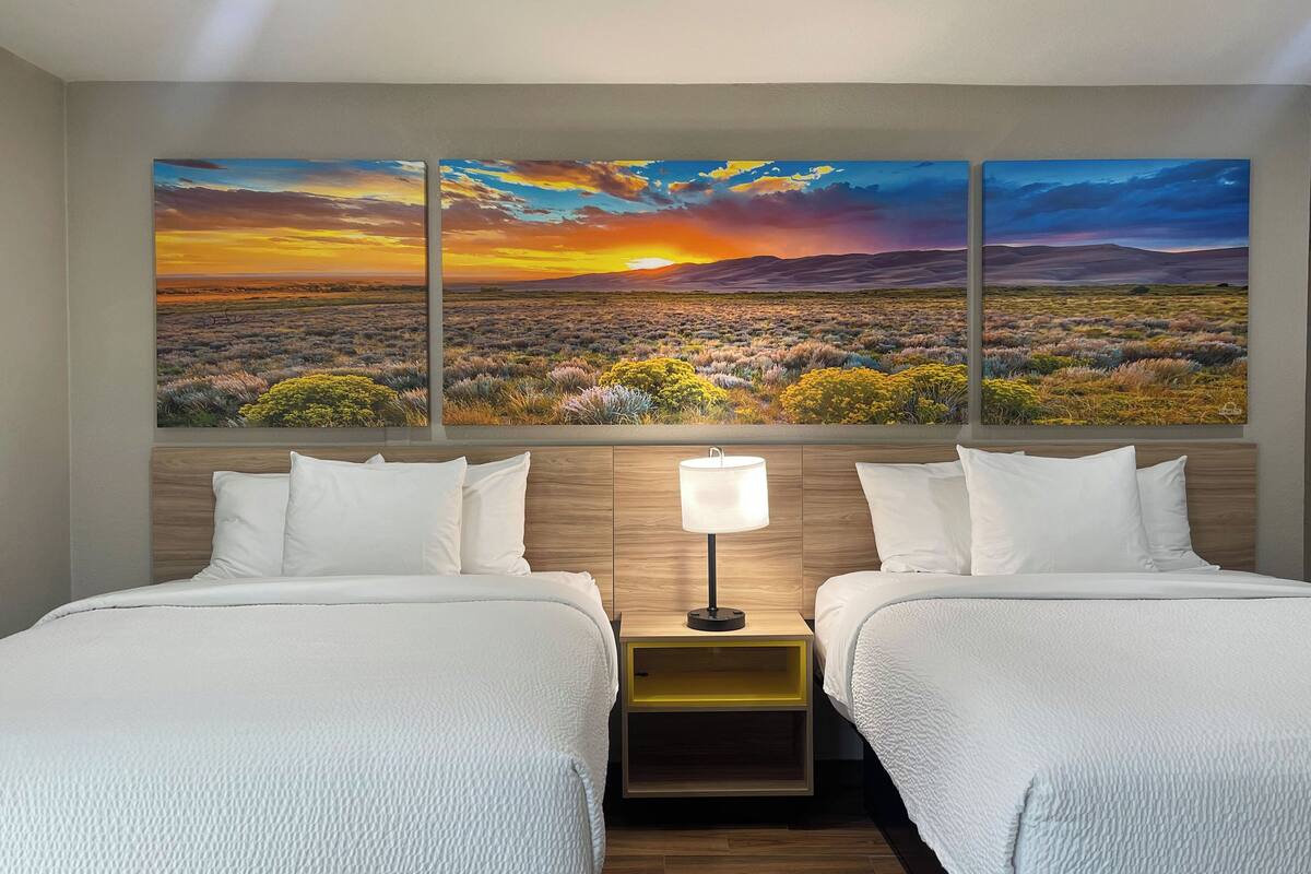 A hotel room with two beds, a nightstand with a lamp, and a large landscape painting above the beds.