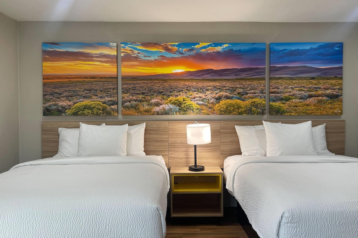 A hotel room with two beds, a nightstand with a lamp, and a wall with three landscape paintings.