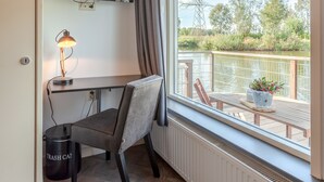 Een kamer met een houten vloer, een bureau met stoel, een aan de muur bevestigde televisie, een raam met uitzicht op een rivier en bomen, en een radiator onder het raam.