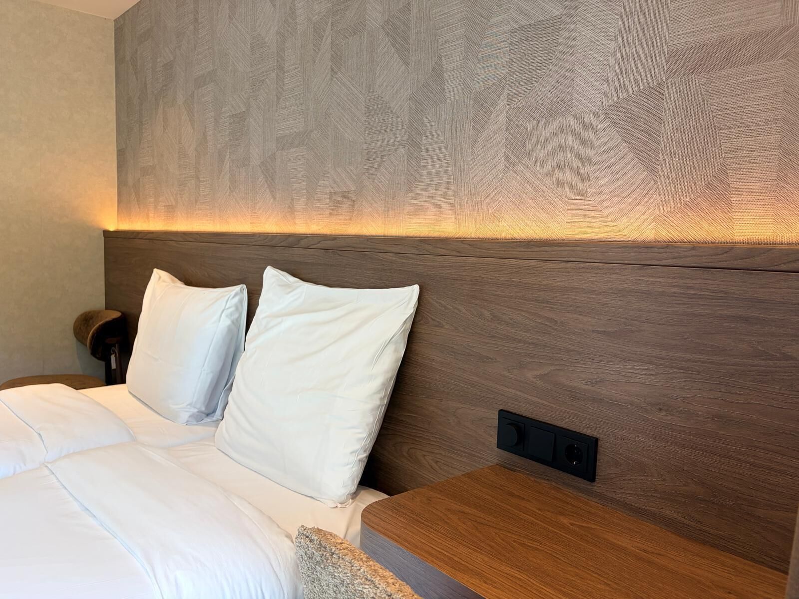 Ein modernes Hotelzimmer mit einem Holzkopfteil, einem Bett mit weißer Bettwäsche und einem Nachttisch.