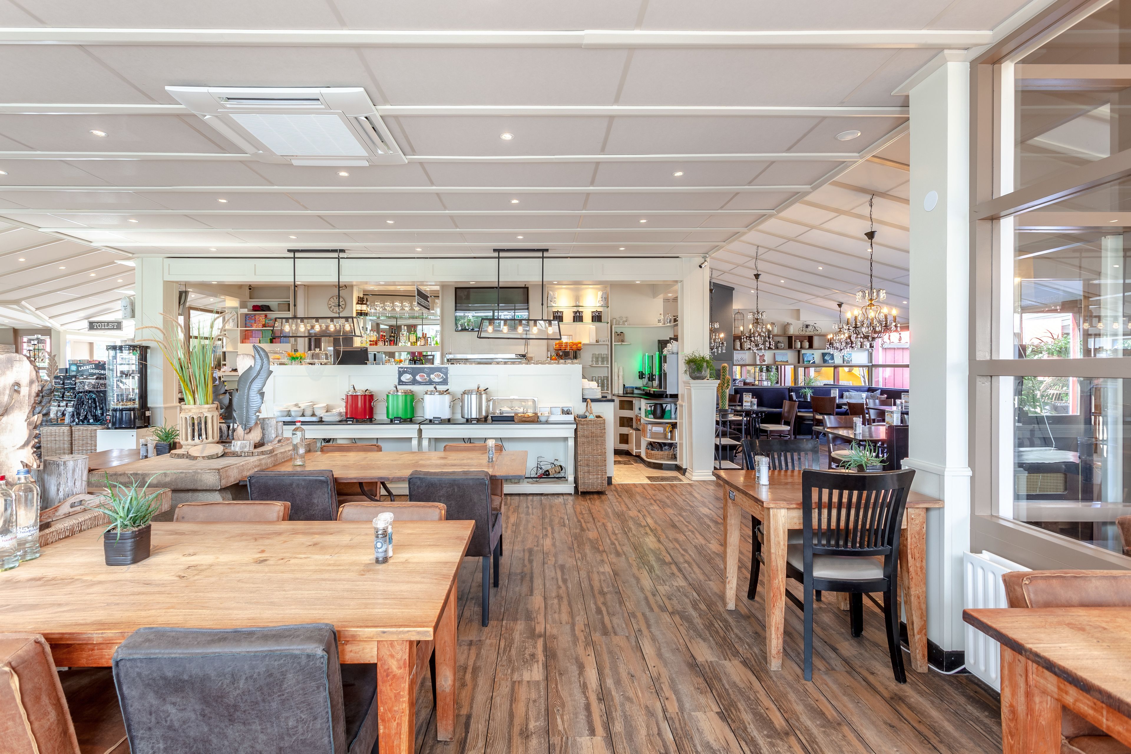 Un restaurante espacioso con piso de madera, una zona central de comedor con mesas y sillas, una cocina al fondo y vistas a través de amplios ventanales.