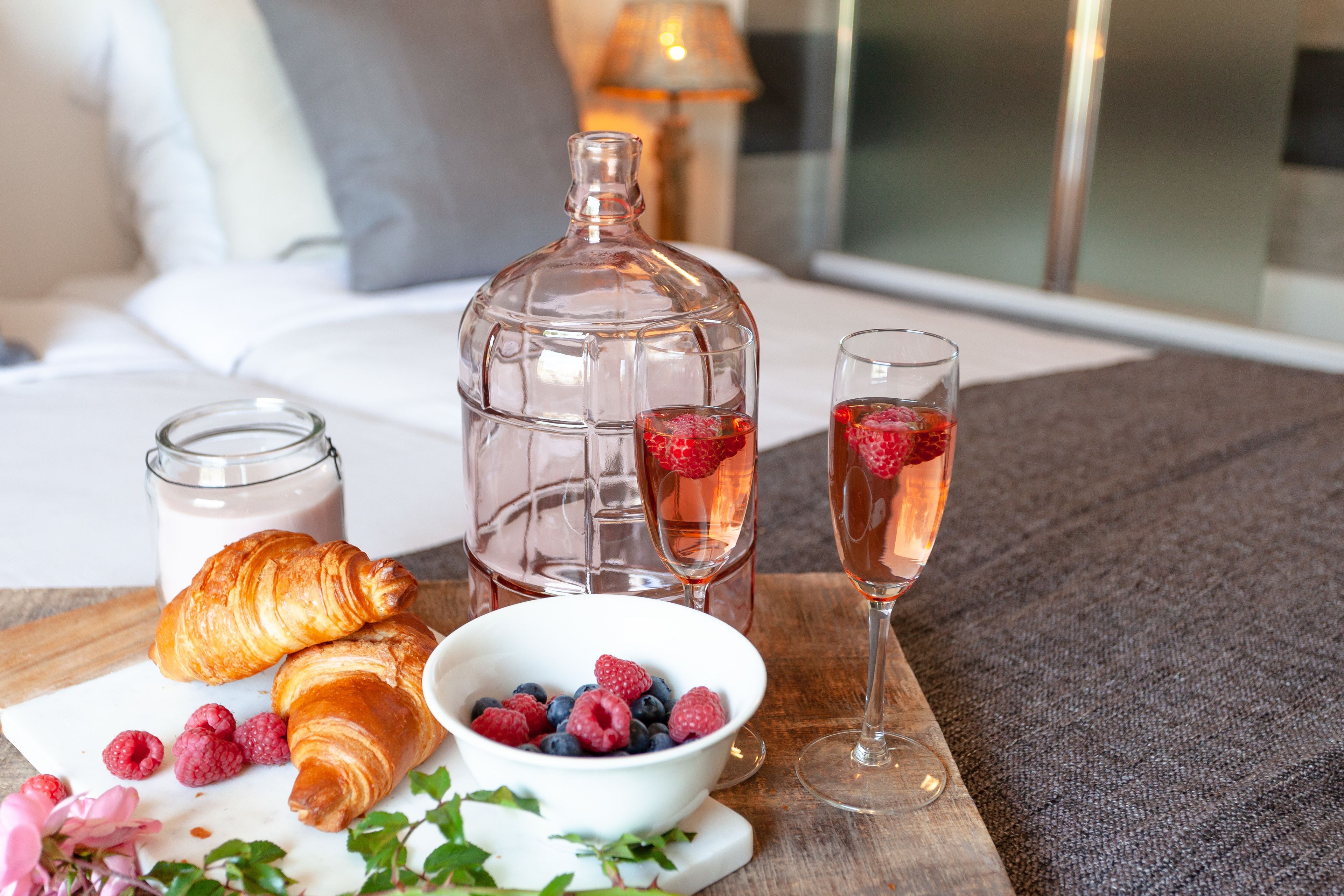 A breakfast setup with croissants, a bowl of berries, a milk jug, and two glasses of a pink beverage on a wooden table.