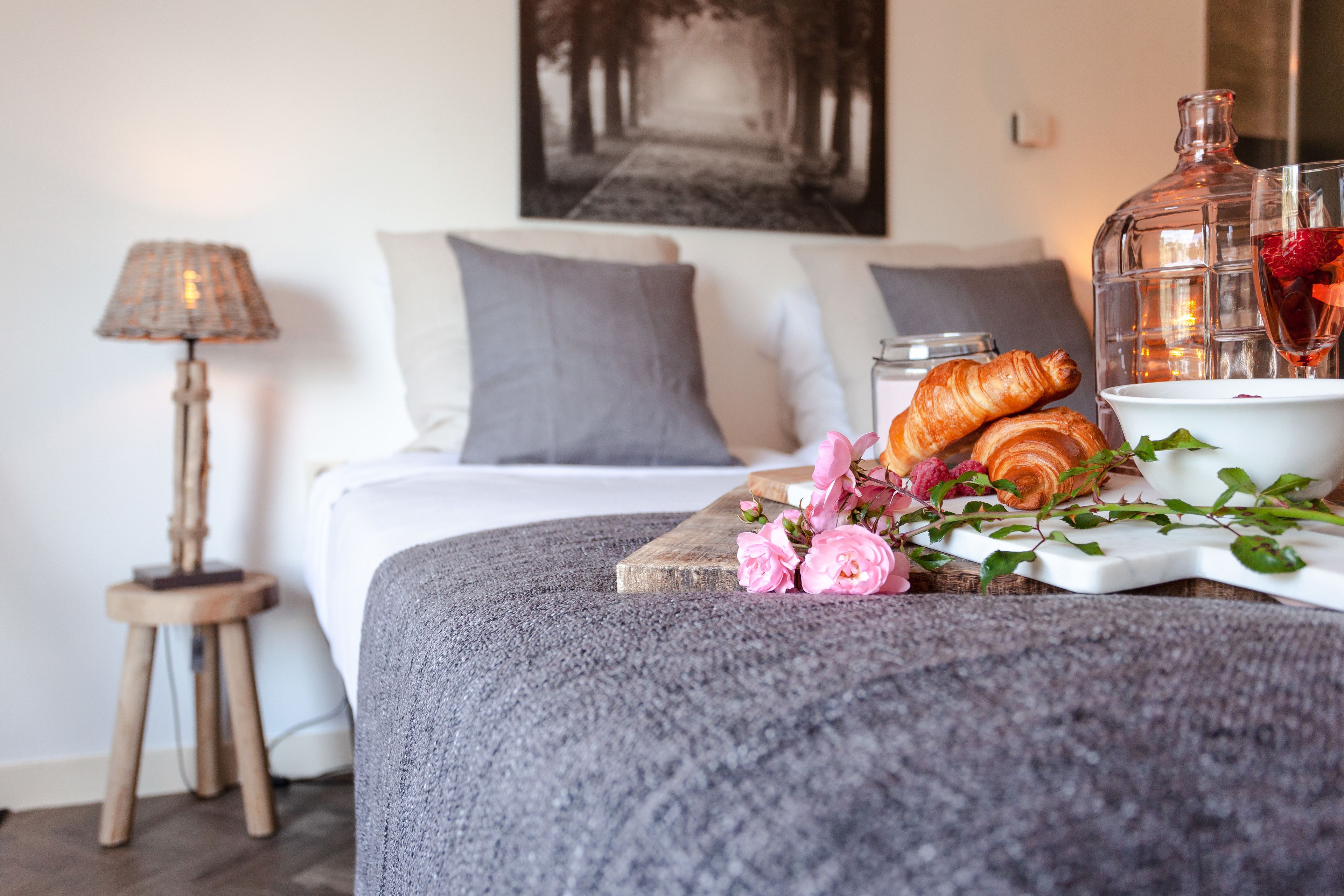 A neatly made bed with pillows, a bedside lamp, a tray with croissants, flowers, and a drink.