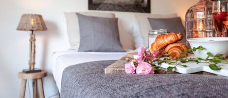 A neatly made bed with pillows, a bedside lamp, a tray with croissants, flowers, and a drink.