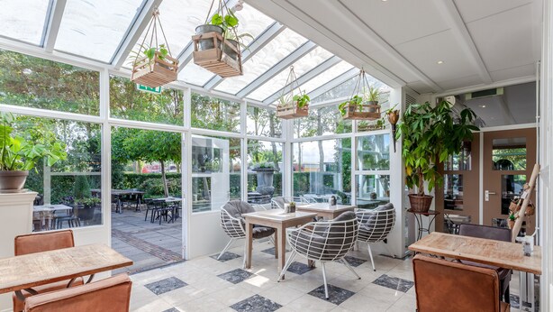 Een ruime conservatorium met grote glazen deuren, houten tafels en stoelen, en hangende plantenbakken.