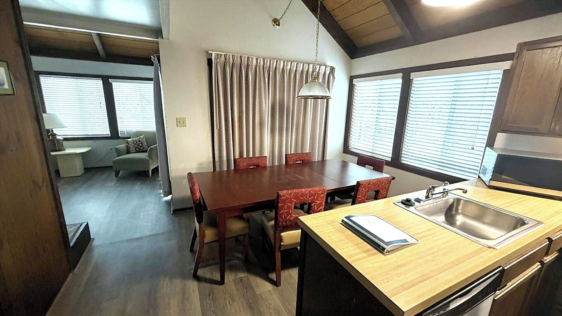 A compact kitchen with a wooden table, four chairs, a hanging light, and a window with blinds.