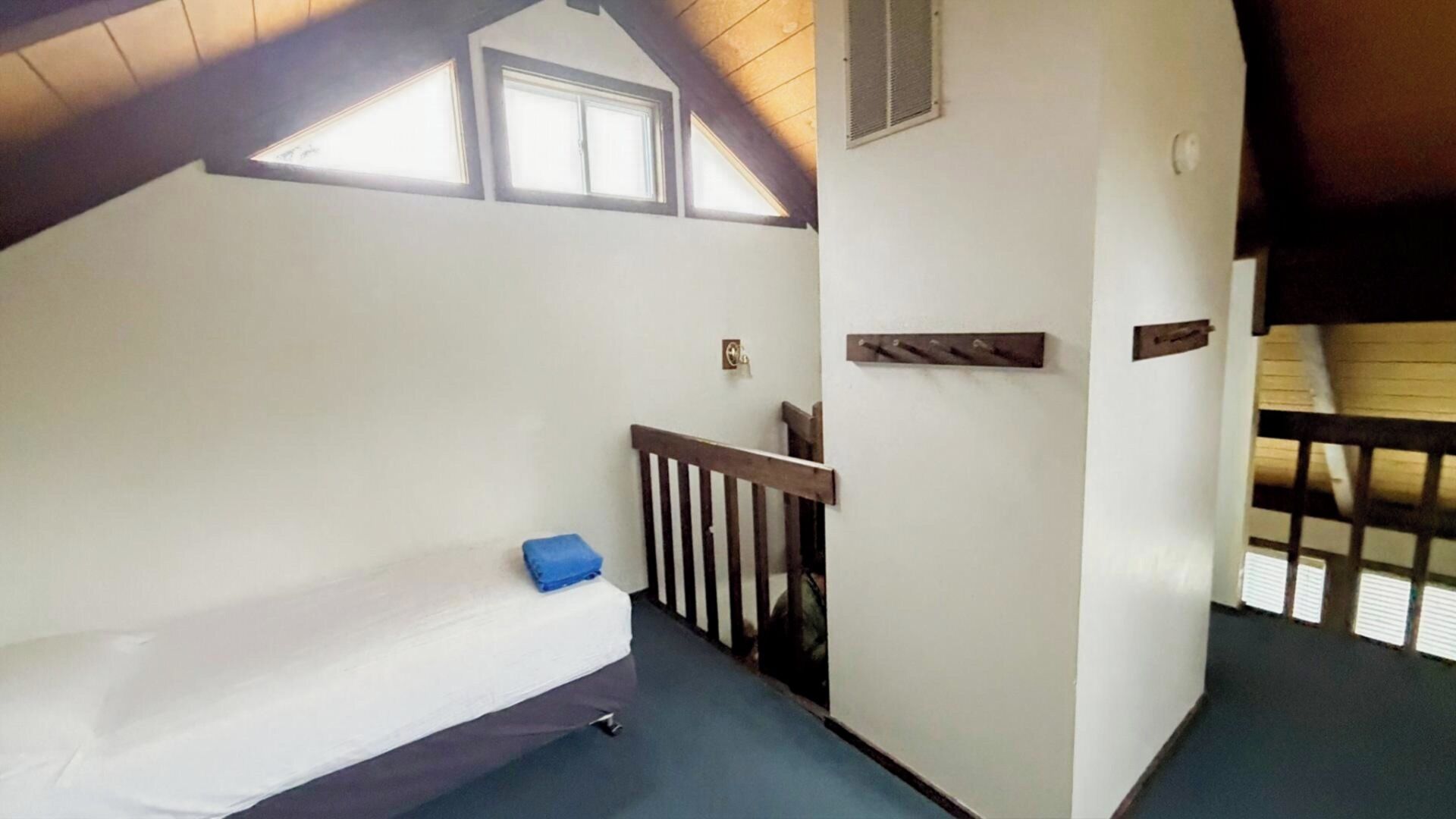 Une petite pièce accueillante avec un plafond en pente, un lit simple avec un oreiller bleu, une rambarde en bois et une petite fenêtre.