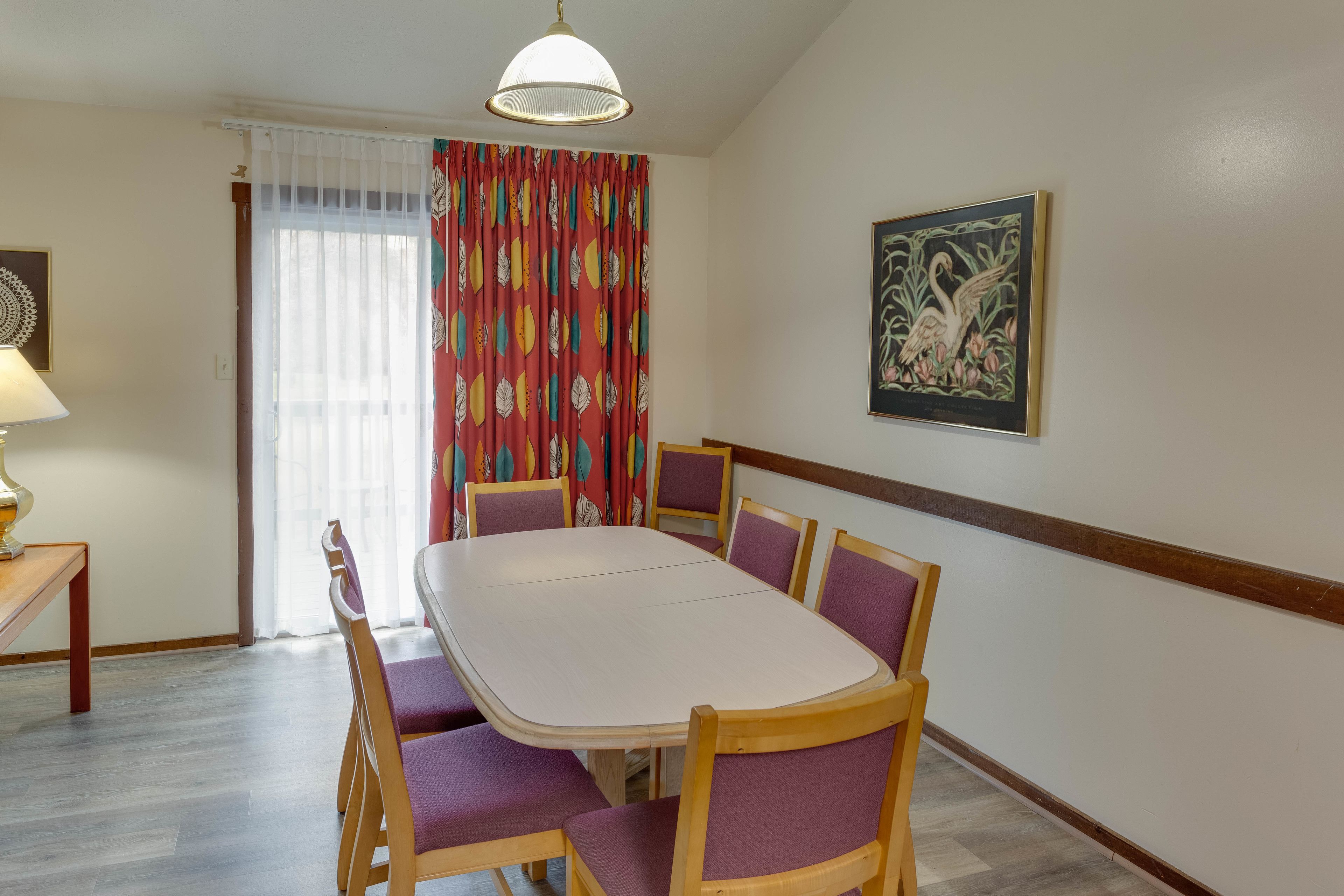 Un coin repas avec une table blanche et des chaises violettes, un luminaire au plafond et une fenêtre avec des rideaux.
