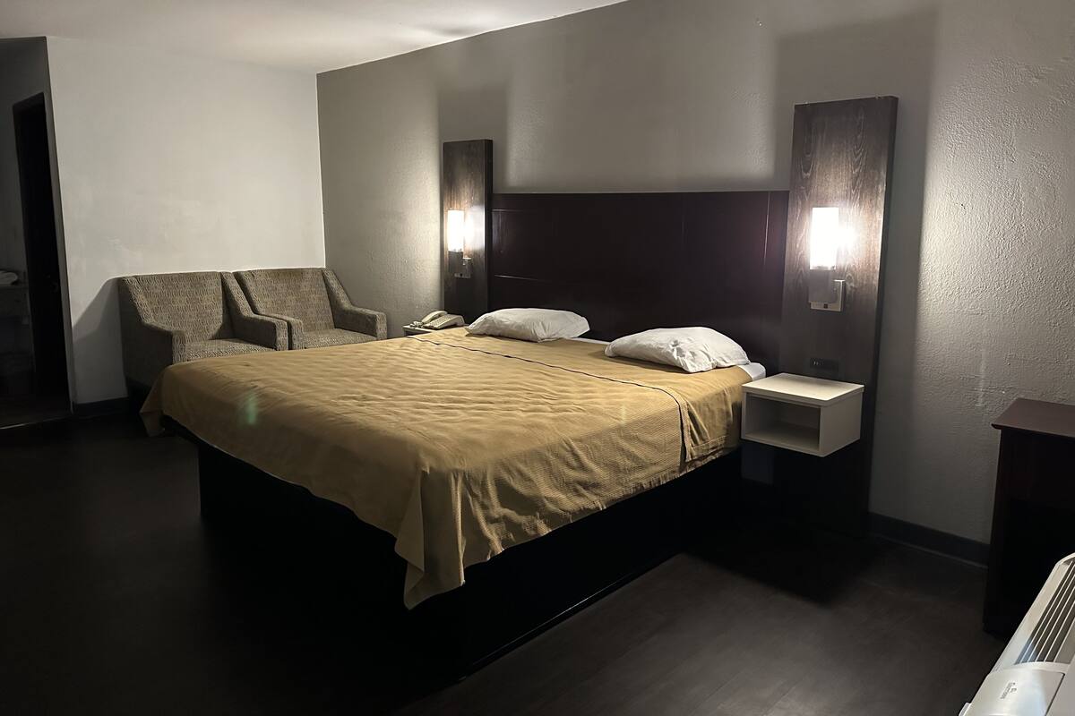 A hotel room with a large bed, a chair, a bedside table, and a wall-mounted light fixture.