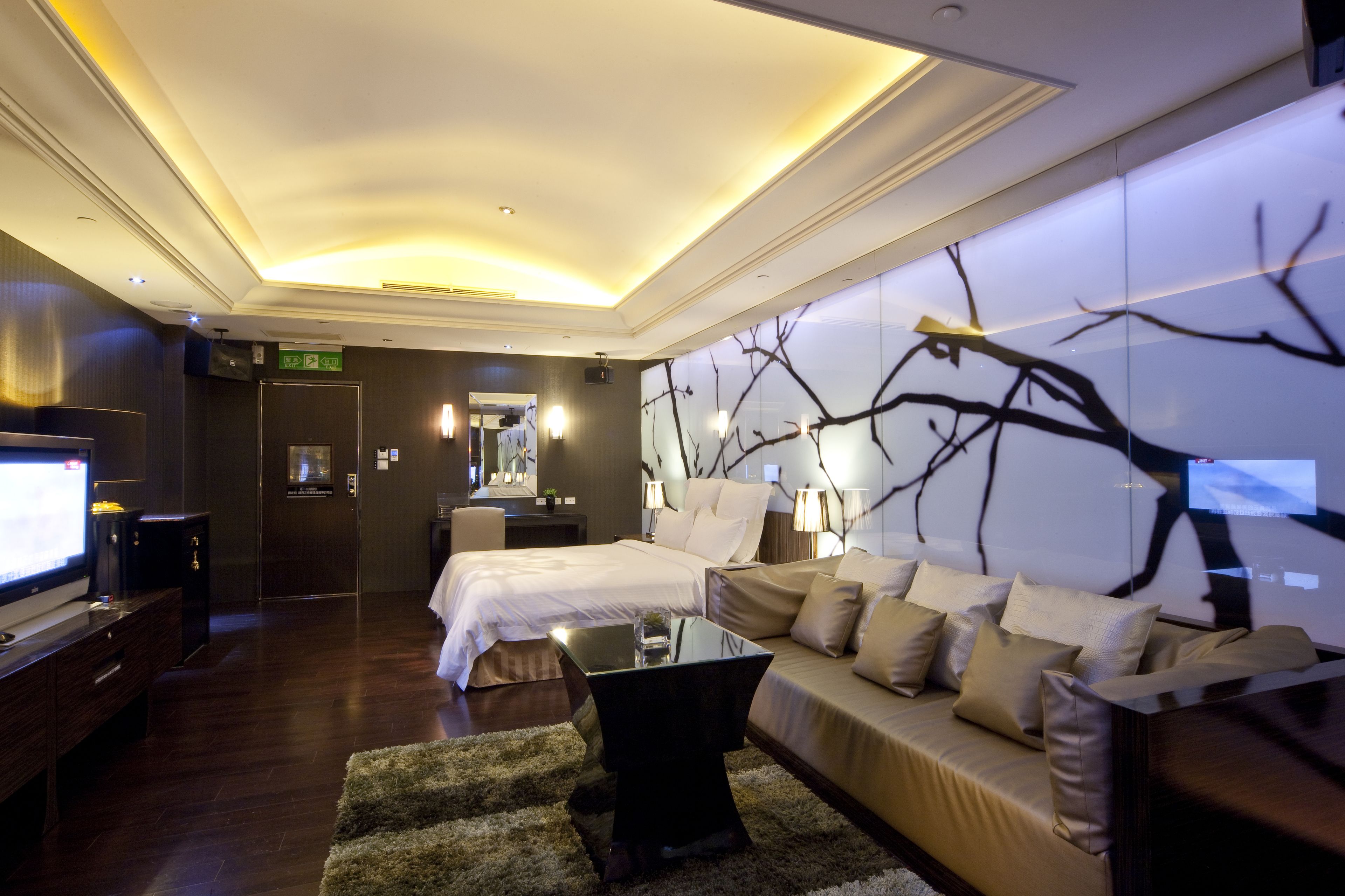 A modern hotel room with a large bed, a sofa, a TV, and a wall with a tree branch design.