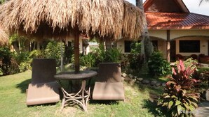 A thatched-roof outdoor seating area with two chairs and a table, surrounded by lush greenery and a building with a red roof. - Aqua-Landia Resort (Dauin)