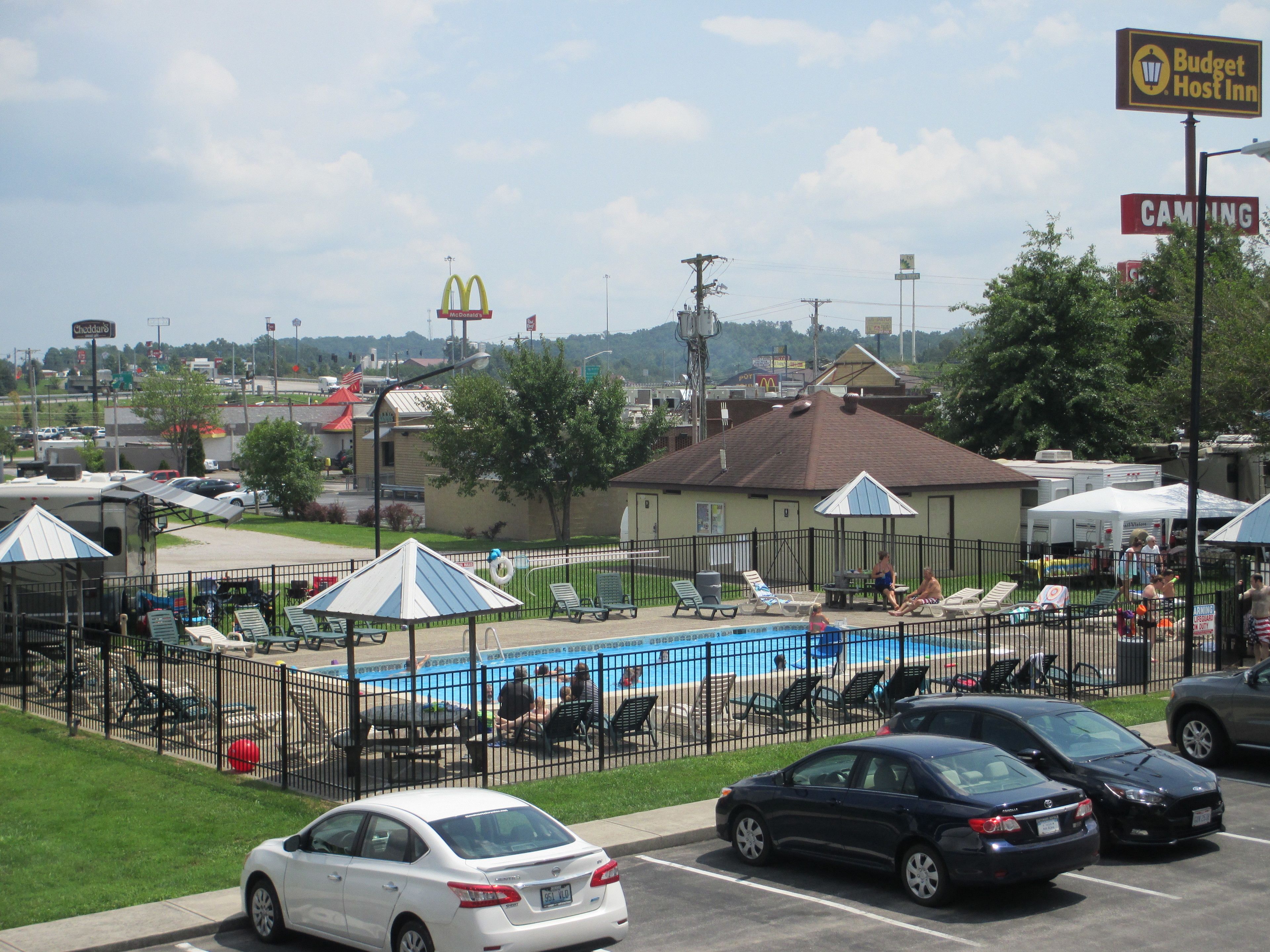 Un espace piscine avec des chaises longues et des parasols, un parking clôturé et un panneau indiquant l’établissement Budget Host Inn.