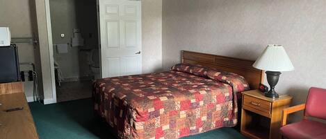A hotel room with a bed, a desk, a chair, a lamp, and a door leading to a bathroom.