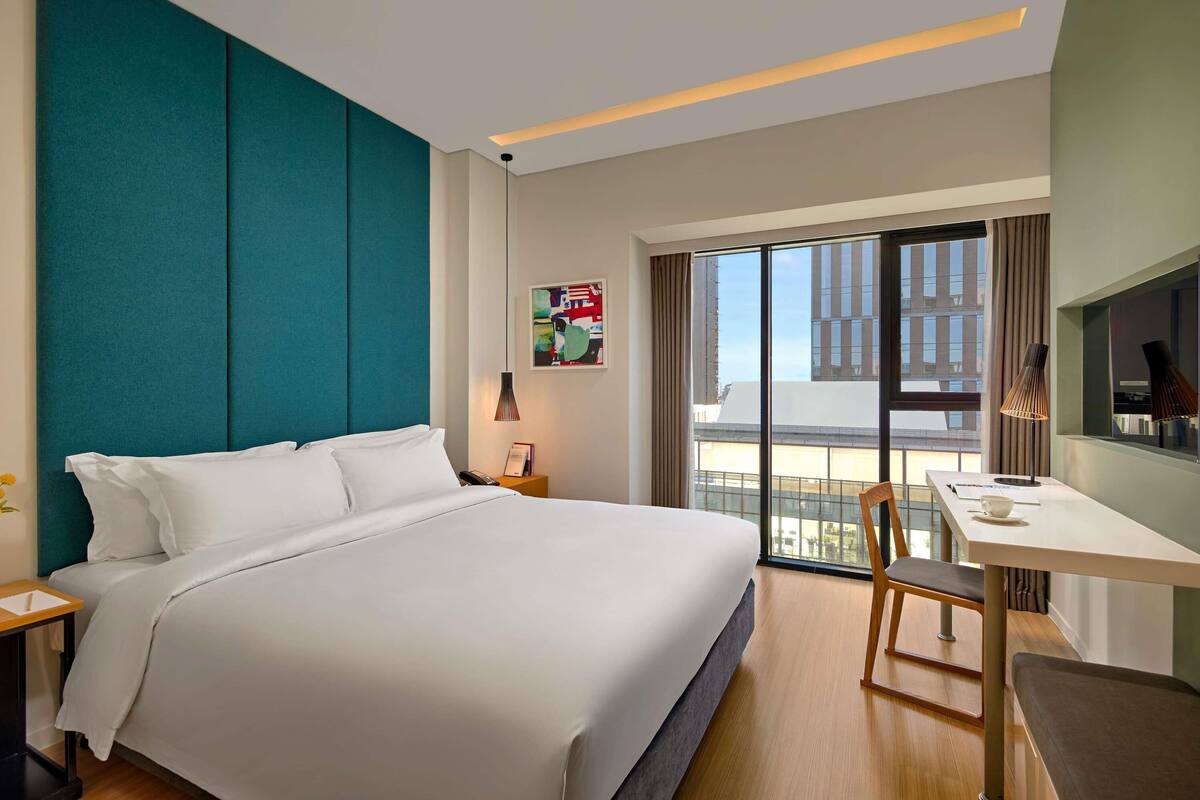 A hotel room with a large bed, a desk, a chair, a nightstand, a lamp, and a view of buildings through the window.