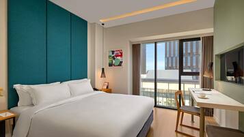Ein Hotelzimmer mit einem großen Bett, einem Schreibtisch, einem Stuhl, einem Nachttisch, einer Lampe und Blick auf Gebäude durch das Fenster.