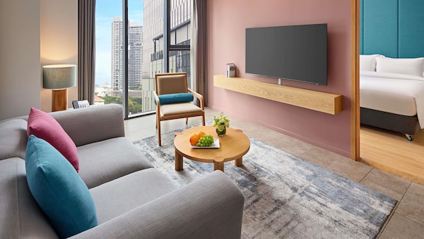 A modern hotel room with a grey sofa, a wooden coffee table with fruit, a TV on a wooden shelf, and a city view through large windows. - Novotel Living Saigon South (Ho Chi Minh City)