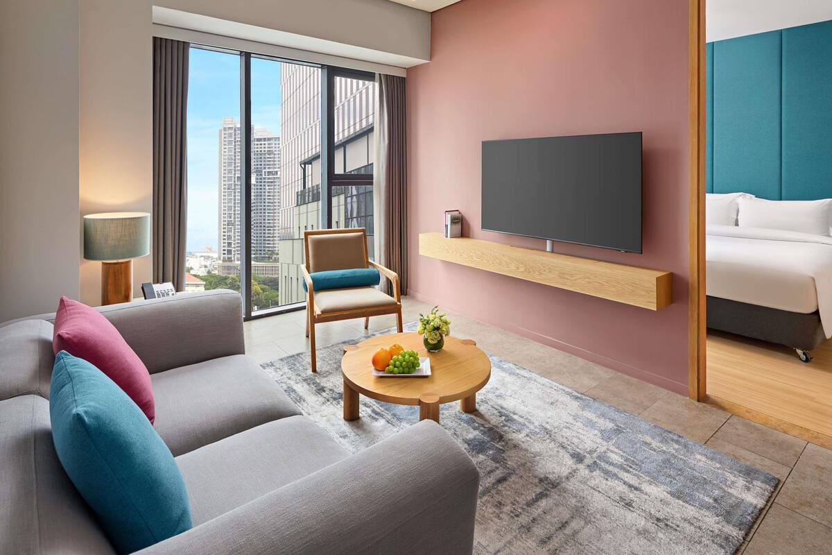 Moderni hotellihuone, jossa on harmaa sohva, puinen sohvapöytä, jossa on hedelmiä, televisio puuhyllyllä ja suuri ikkuna, josta avautuu näkymä kaupunkiin.
