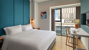A modern hotel room with a large bed, a desk with a chair, a window with a view of buildings, and a painting on the wall. - Novotel Living Saigon South (Ho Chi Minh City)