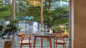 A dining area with a table set for two, a large window with a view of trees and buildings, and a hanging light fixture. - Novotel Living Saigon South (Ho Chi Minh City)