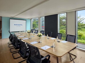 A conference room with a long table, chairs, a projector screen, and a view of greenery outside. - Novotel Living Saigon South (Ho Chi Minh City)
