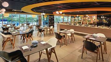 A modern restaurant with multiple dining tables and chairs, a counter with food display, and large windows offering a view of greenery.
