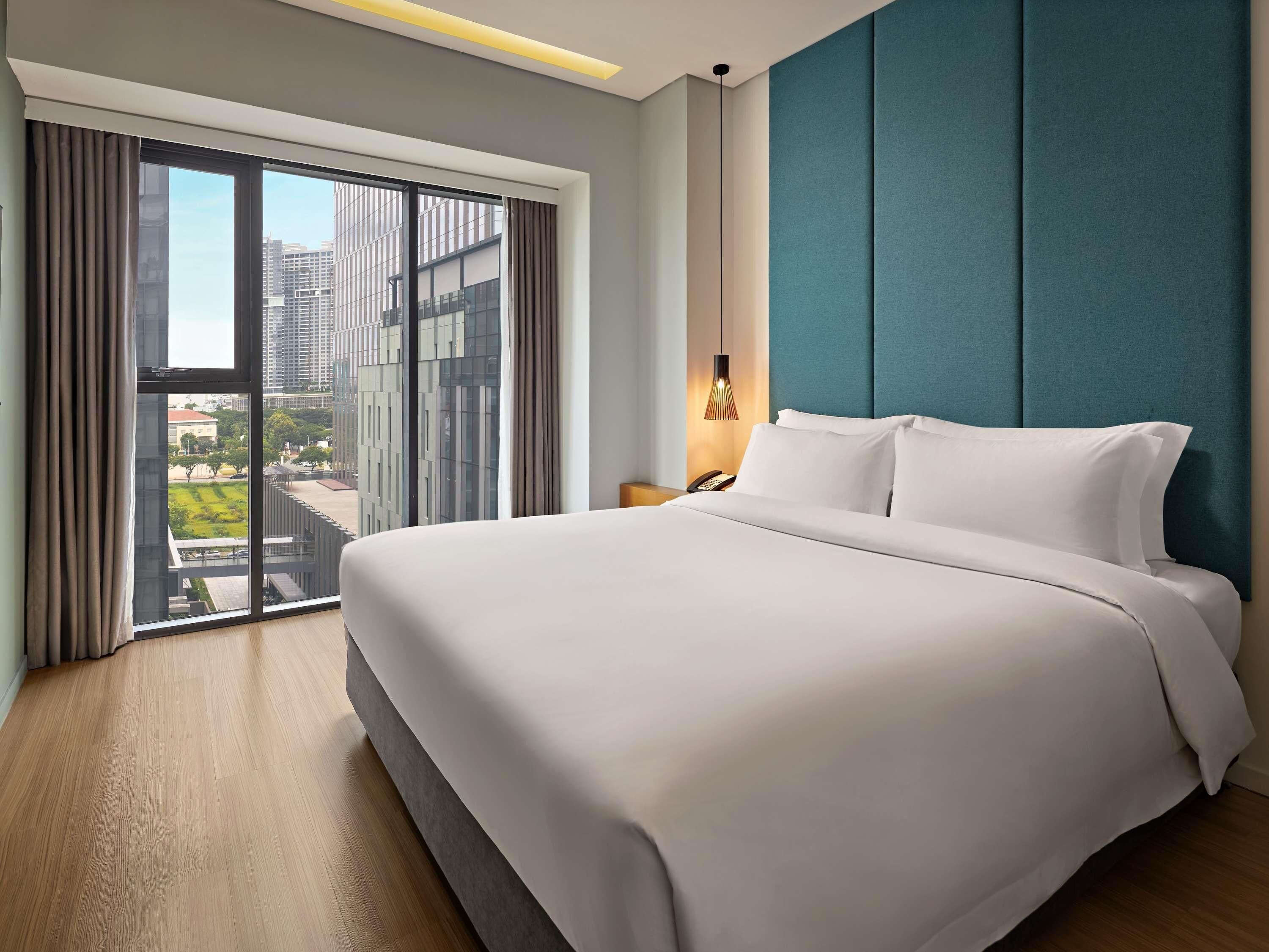 A modern hotel room with a large bed, a view of the city through floor-to-ceiling windows, and a cityscape visible outside.