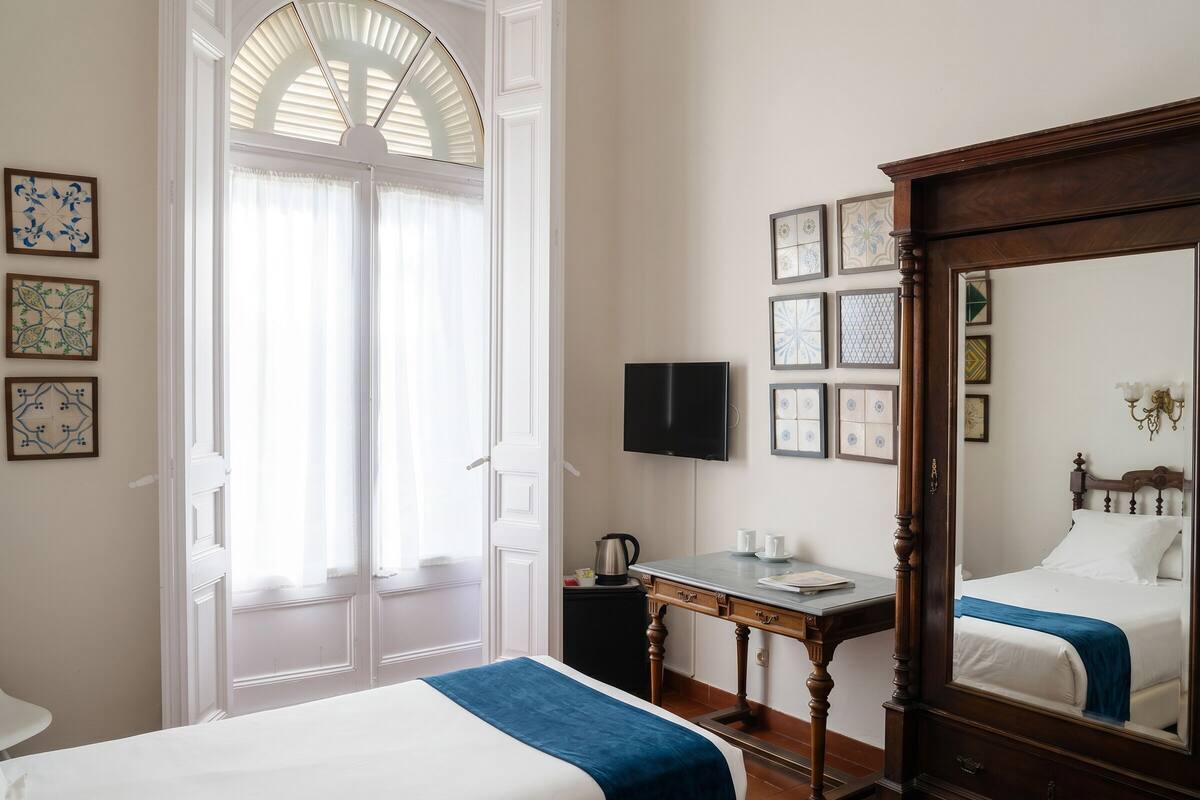 Habitación de hotel con cama, escritorio con silla, televisor, espejo grande y ventana con cortinas.