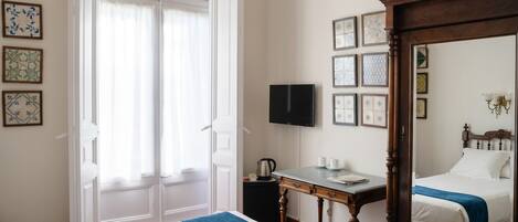 Une chambre d’hôtel comprenant un lit, un bureau avec une chaise, une télévision, un grand miroir et une fenêtre avec des rideaux.