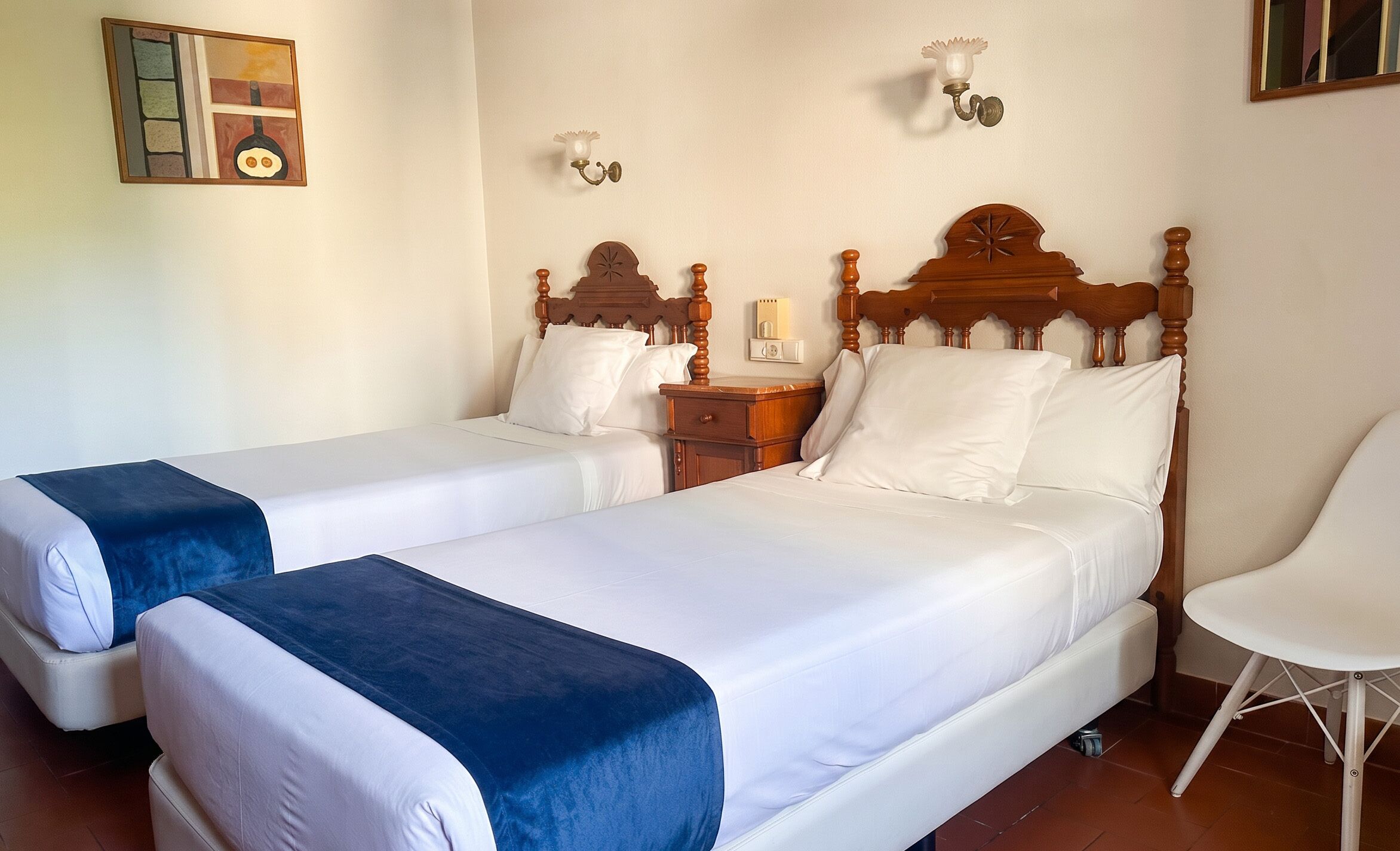 Habitació d'hotel amb dos llits, una tauleta de nit, una cadira i una làmpada penjada a la paret.