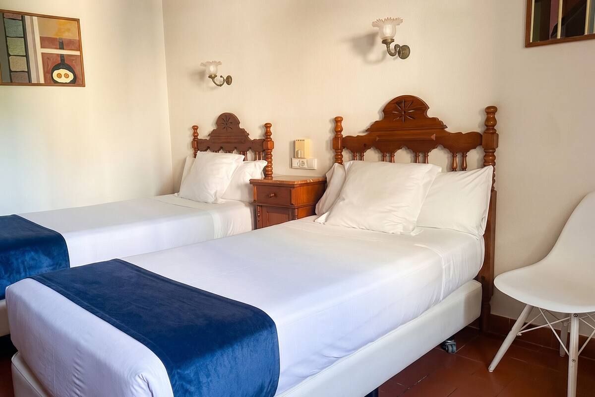 Habitación de hotel con dos camas, una mesita de noche, una silla y una lámpara fijada a la pared.