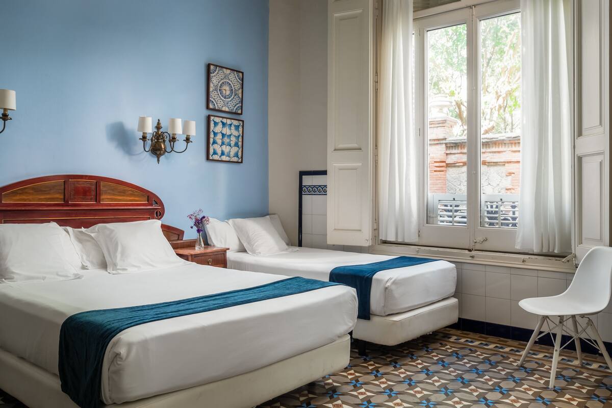 Una habitación de hotel con dos camas, una ventana con cortinas, piso estampado y una silla blanca.