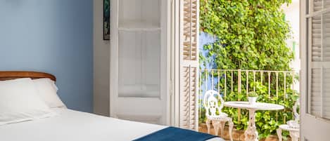 Un dormitorio con cama, una puerta blanca y un balcón con barandilla blanca y plantas verdes.