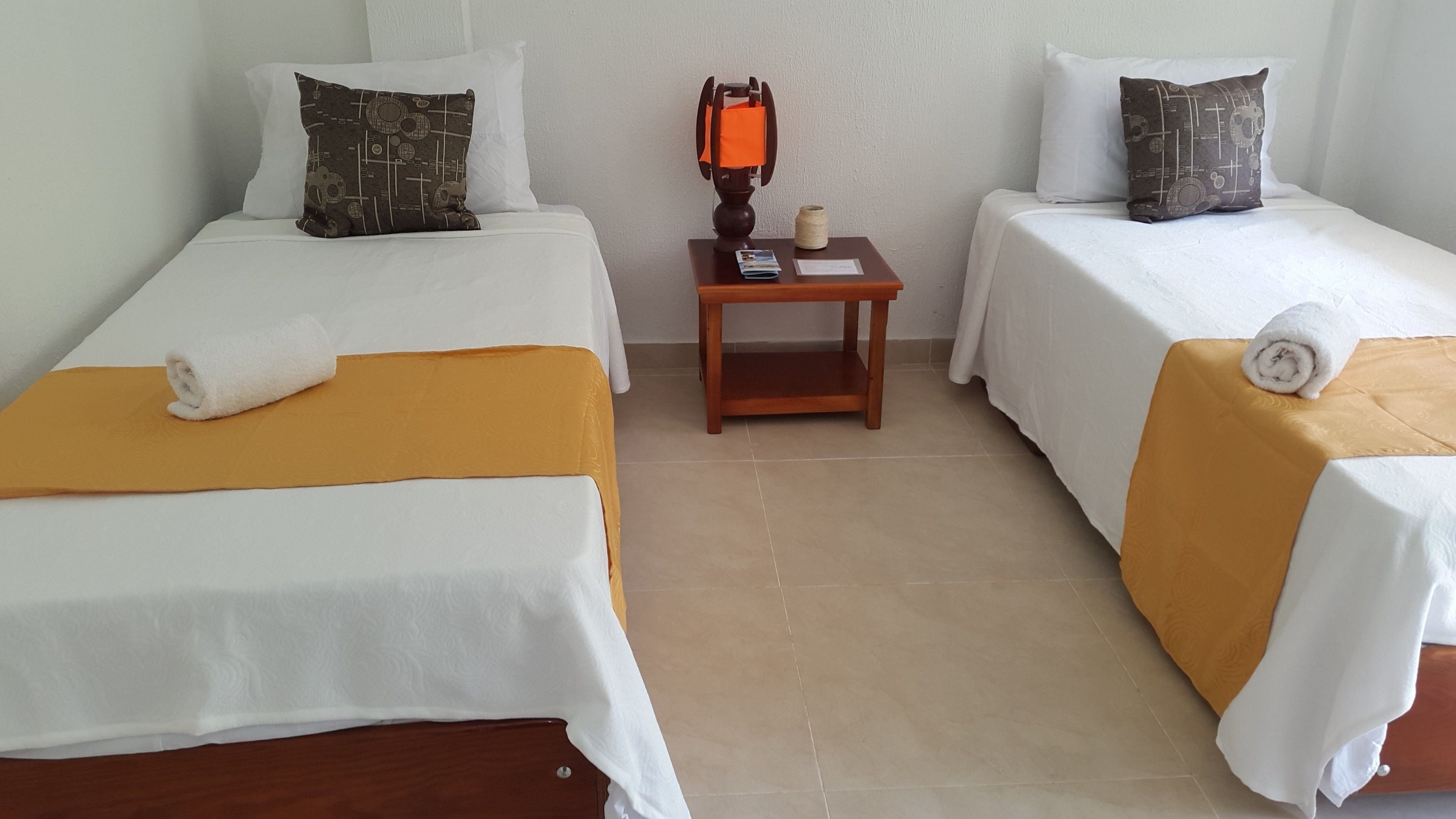 Une chambre d’hôtel avec deux lits simples, chacun recouvert d’une courtepointe blanche et agrémenté d’un oreiller marron. Une petite table de chevet en bois est placée entre les lits, sur laquelle se trouvent une bobine de fil et un livre.
