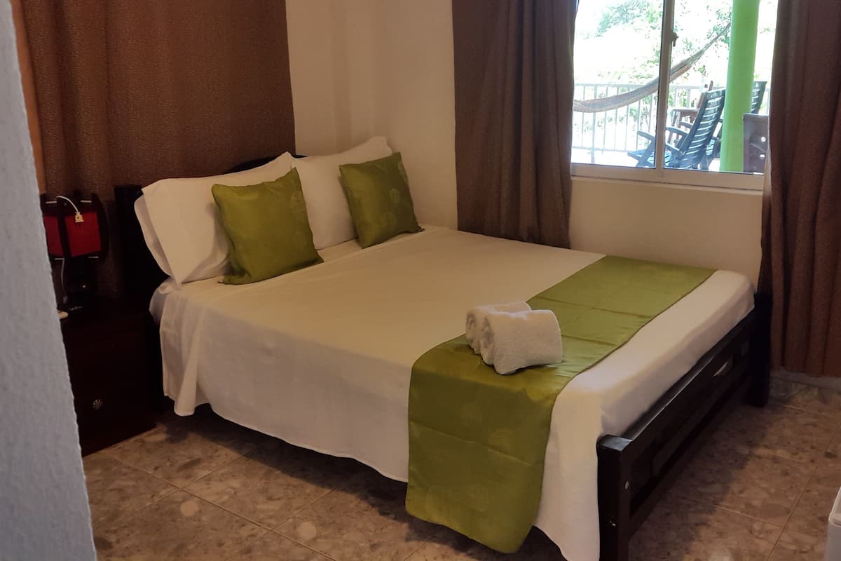 Una cama bien hecha con una colcha blanca y verde, un almohadar blanco y un almohadar verde. Hay una toalla blanca sobre la cama. La habitación tiene una ventana con cortinas marrones, un suelo de baldosas y un radiador blanco en el suelo.