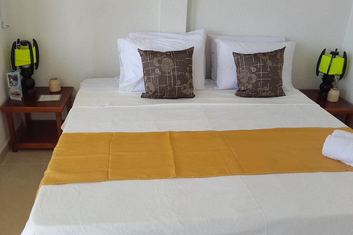 Una cama bien hecha con una manta amarilla, almohadas blancas y una toalla blanca a un lado. Hay dos mesitas de noche, cada una con una lámpara y un pequeño florero.