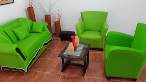 Un canapé vert, deux fauteuils verts, une petite table avec un vase, et de grands vases ornés de branches rouges, le tout placé devant un mur blanc.