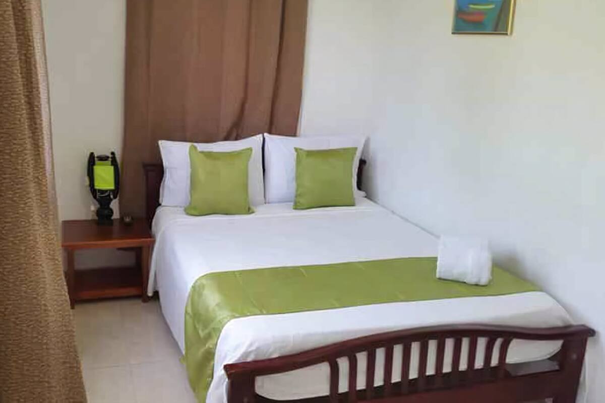Una cama bien hecha con una colcha verde y blanca, un cabecero de madera y una mesita de noche con una lámpara.
