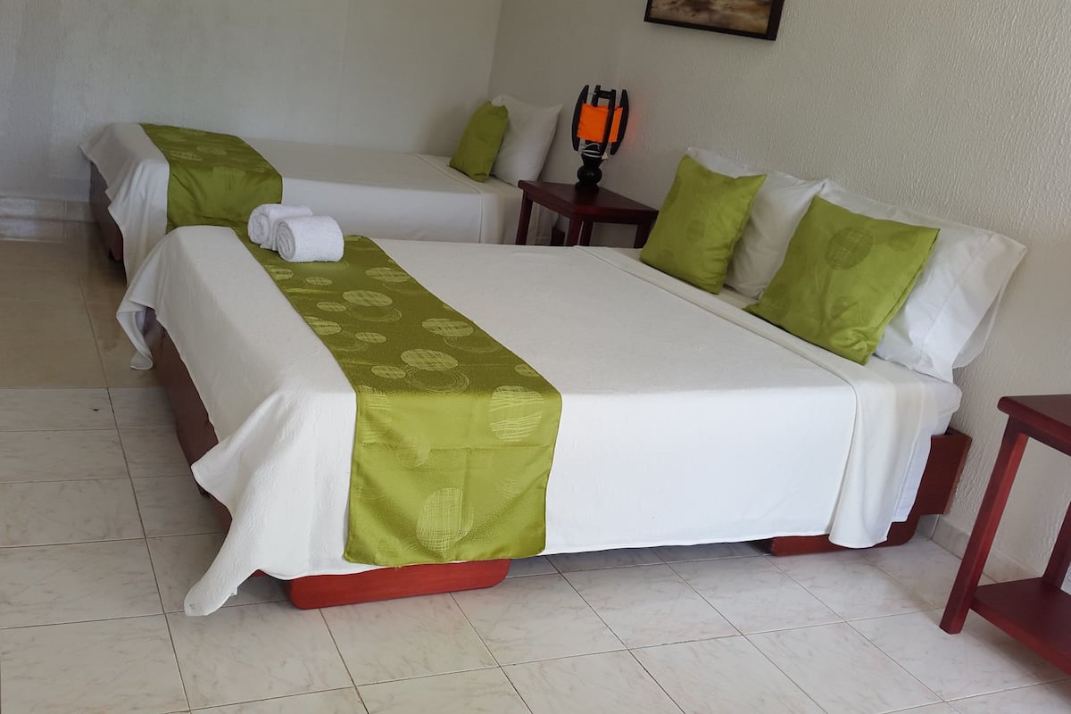 Habitación de hotel con dos camas, cada una con ropa blanca y detalles verdes, una mesita con lámpara y piso de baldosas.