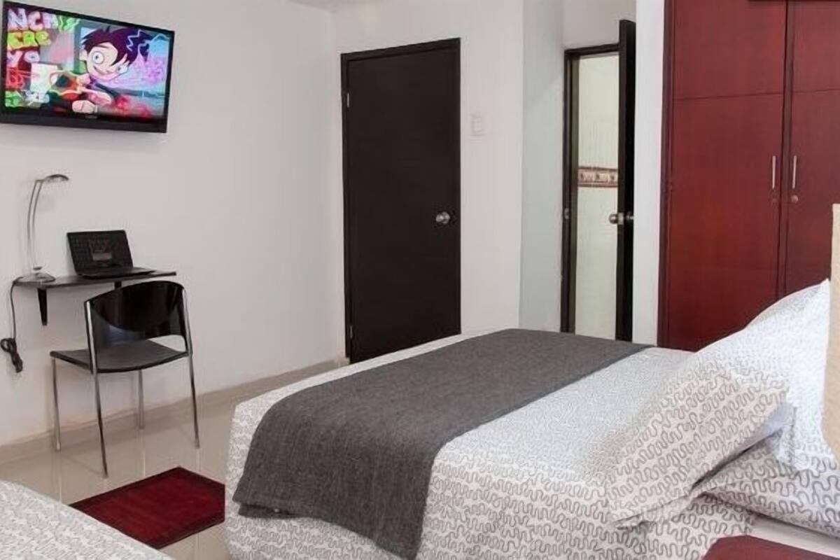 A bedroom with a bed, a desk with a laptop, a chair, a TV on the wall, and a wardrobe.