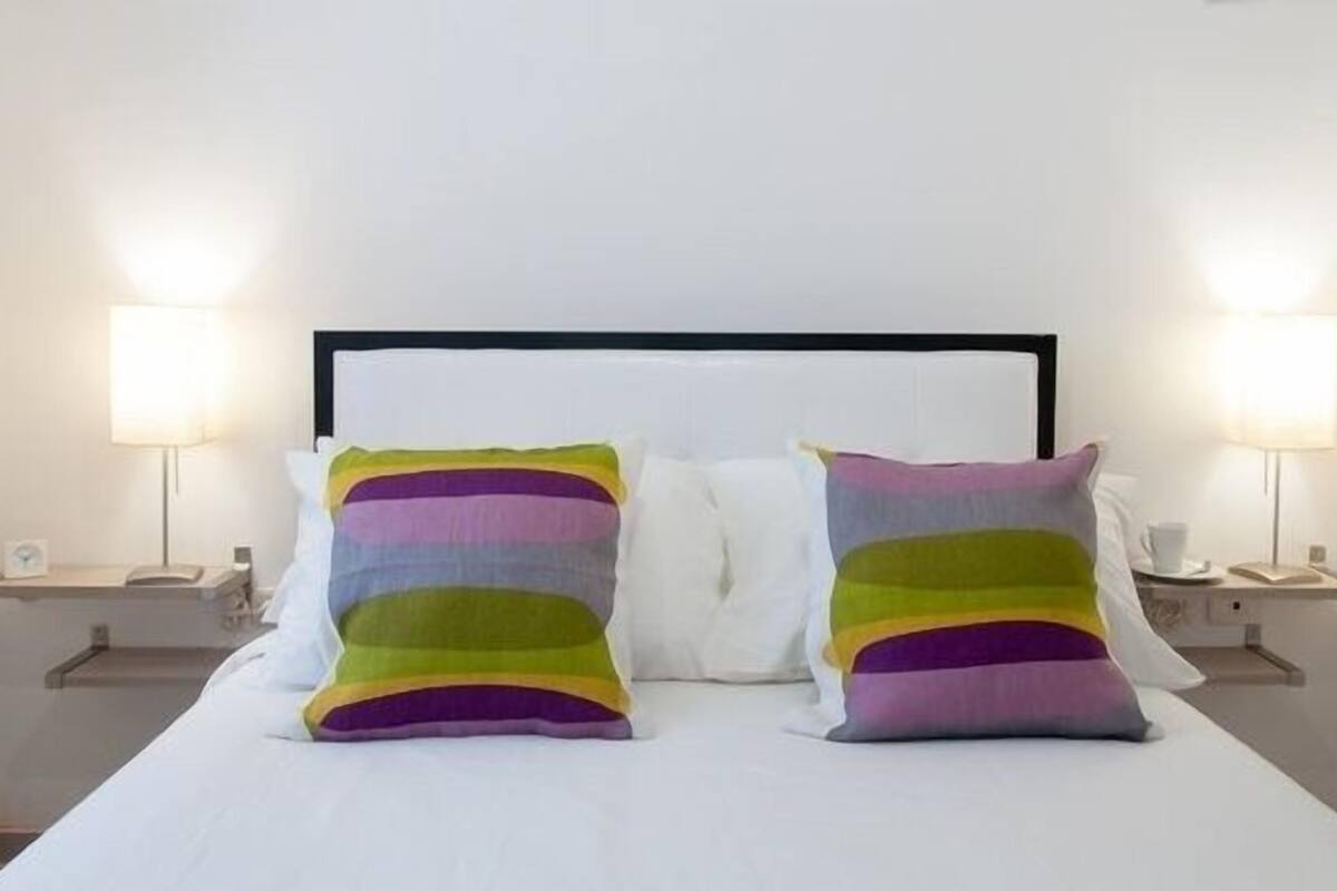 A bed with white linens, two pillows with multicolored stripes, and two bedside lamps on nightstands.