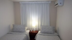 A hotel room with two single beds, a nightstand with a lamp, and a window with white curtains.